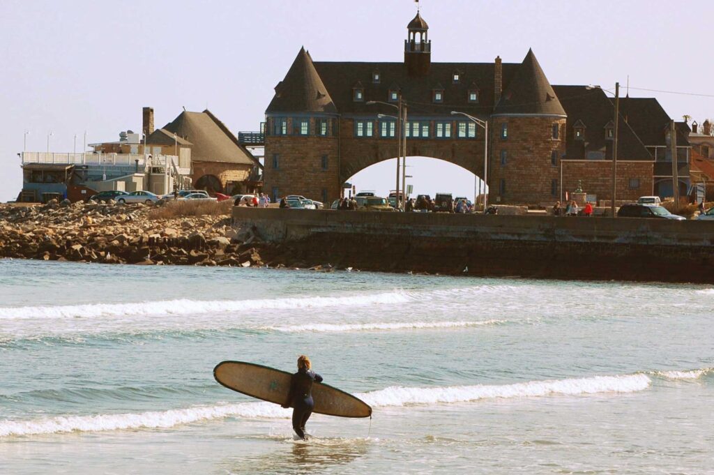 A surfer standing in the water with the Narragansett towers in the background